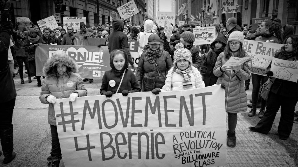 black and white photo of marchers holding a banner that reads "#movement4bernie"